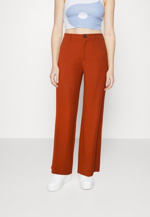Femme portant un pantalon large taille haute couleur rouille, des baskets à plateforme blanches, et un top court sans manches bleu clair et blanc.