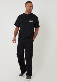 Hombre con camiseta negra con gráfico, pantalones cargo negros y zapatillas grises, de pie con una mano en el bolsillo y sonriendo.