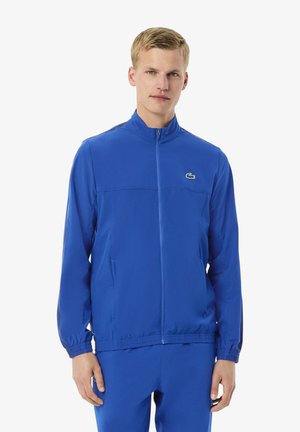 Joven vestido con una chaqueta deportiva azul con cremallera y pantalones a juego, de pie contra un fondo blanco liso.