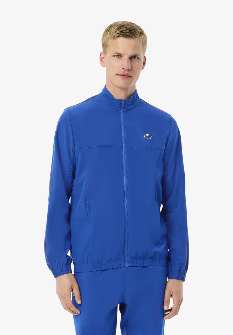 Junger Mann in einer blauen Sportjacke mit Reißverschluss und passenden Hosen, der vor einem schlichten weißen Hintergrund steht.