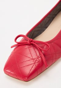 Ballerine en cuir rouge avec un bout carré, présentant un motif matelassé et un petit nœud sur la tige. Texture lisse, design léger.
