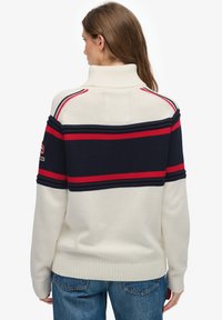 Pull en maille crème, avec des rayures horizontales bleu marine et rouges. Col montant et poignets côtelés ajoutent de la texture. Coutures détaillées sur les épaules.