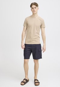 Beige bomull t-skjorte med rund hals, parret med mørk marineblå shorts med sidelommer og avslappet passform. Brune sandaler fullfører antrekket.