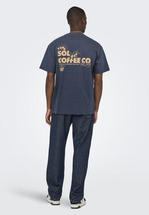 Man draagt een marineblauw T-shirt met de tekst "Sol Coffee Co" en een beige bloemenpatroon, gecombineerd met donkerblauwe spijkerbroek, staand tegen een effen lichte achtergrond.