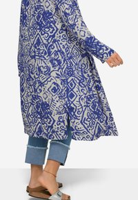 Lange, lichtgewicht tuniek in crème met blauwe abstracte patronen. Beschikt over lange mouwen en zijsplitten. Gepaard met omgeslagen denim en sandalen.