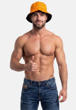 Muskularny mężczyzna bez koszulki, noszący pomarańczowy kapelusz typu bucket z abstrakcyjną grafiką linii, pokazujący kciuk w górę, ubrany w niebieskie dżinsy na jednolitym tle.