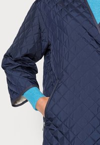 Giacca trapuntata blu navy con motivo a diamante, caratterizzata da una cerniera frontale e maniche oversize. I polsini interni di un blu chiaro aggiungono un dettaglio a contrasto.