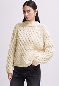 Pull en crème en maille avec un motif en nid d'abeille texturé, col montant à côtes hautes et manches longues, assorti à un jean foncé et une bague en argent.
