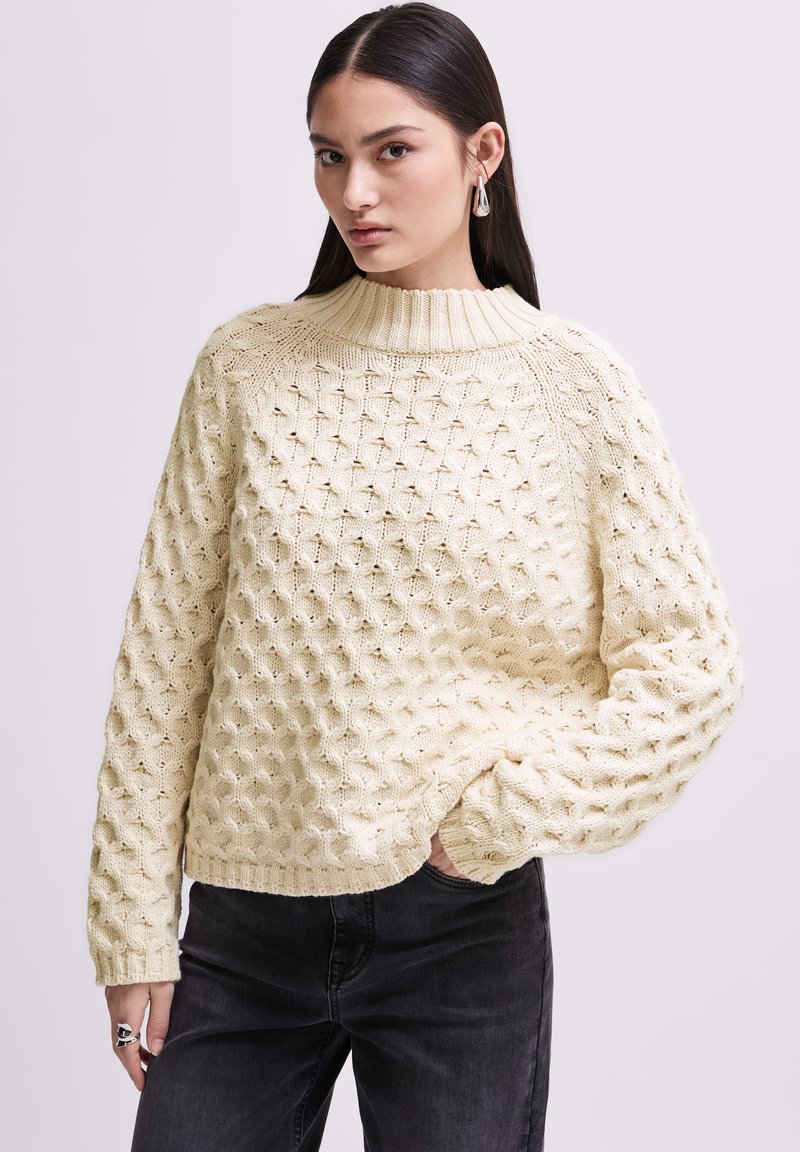 Pull en crème en maille avec un motif en nid d'abeille texturé, col montant à côtes hautes et manches longues, assorti à un jean foncé et une bague en argent.