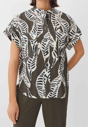 Vrouw draagt een blouse met korte mouwen en knopen, met een zwart-wit bladerenpatroon en twee borstzakken aan de voorkant, gecombineerd met een donkere broek.