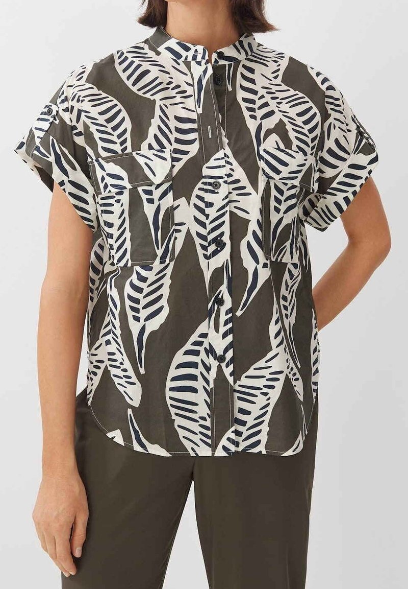 Femme portant une chemise à manches courtes boutonnée avec un motif de feuilles noir et blanc et deux poches poitrine avant, associée à un pantalon sombre.