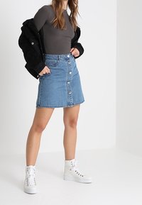 Denimrock in Hellblau mit Knopfdetails auf der Vorderseite, kombiniert mit einer dunklen Jacke, einem grauen Oberteil und weißen High-Top-Sneakers.