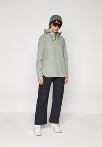 Giacca impermeabile verde chiaro con cappuccio e cordini regolabili, abbinata a pantaloni cargo neri e sneakers beige.