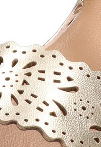 Detalle recortado en metal dorado en una sandalia beige, que muestra intrincados patrones florales y acentos texturizados a lo largo del borde.