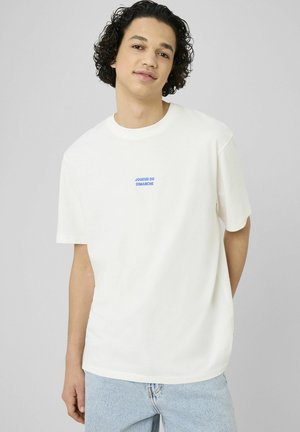 Jeune homme aux cheveux bouclés foncés portant un t-shirt blanc avec un texte bleu « JOUEUR DU DIMANCHE » et un jean bleu clair sur un fond uni.