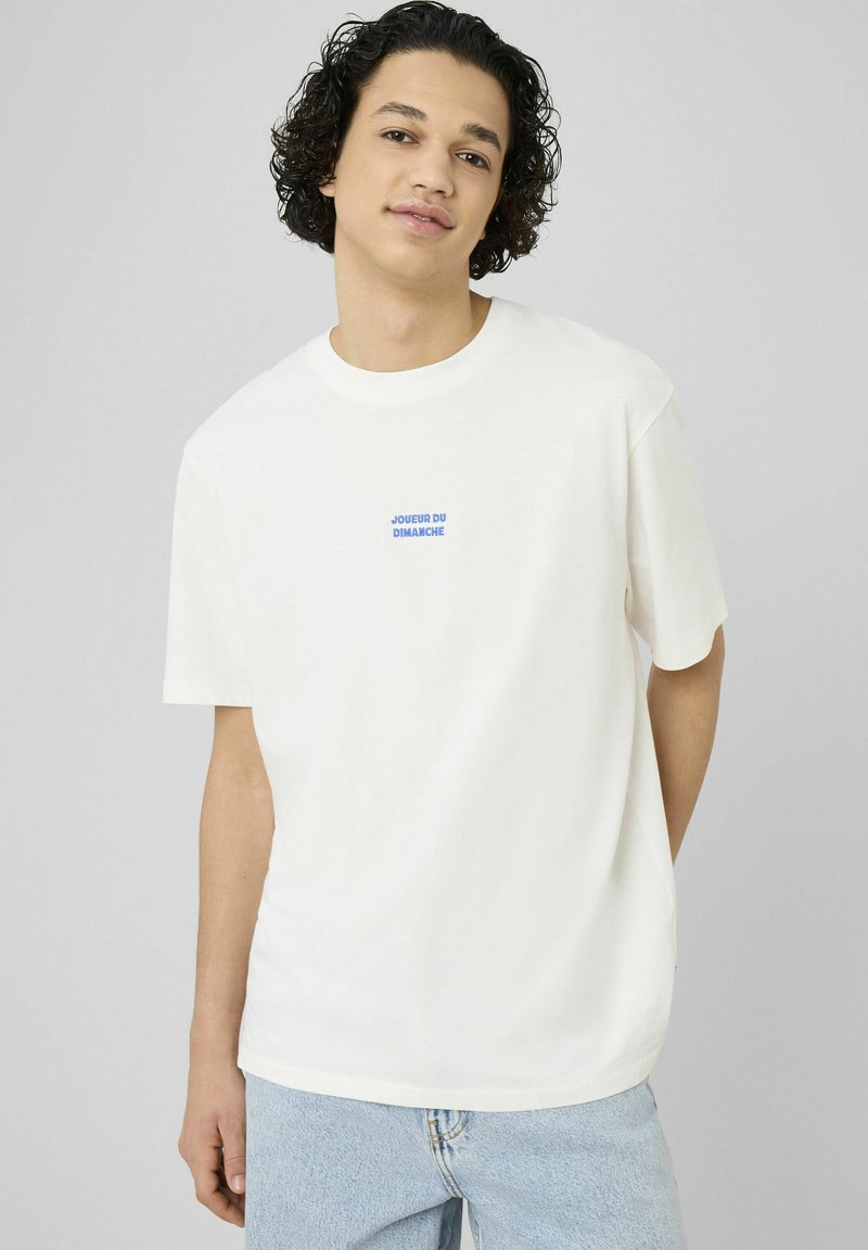 Jeune homme aux cheveux bouclés foncés portant un t-shirt blanc avec un texte bleu « JOUEUR DU DIMANCHE » et un jean bleu clair sur un fond uni.