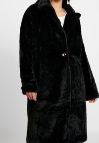 Musta tekoturkivatakki pehmeällä pinnalla, ylisuuri malli, leveä kaulus ja yksittäinen napin kiinnitys. Sisältää pitkät hihat ja sileän pinnan.