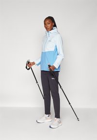 Veste imperméable bleu clair et blanche avec capuche, associée à un pantalon sombre et des chaussures de sport grises, tenant des bâtons de randonnée.