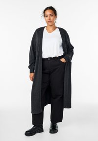 Langer, dunkelgrauer Cardigan aus weichem Stoff, getragen über einem weißen T-Shirt und schwarzen Hosen. Hat eine lockere Passform und seitliche Taschen. Schwarze Stiefeletten.