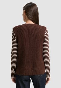 Frau mit glattem braunem Haar, die einen ärmellosen, grob gestrickten braunen Pullunder über einem langärmeligen, braun-weißen gestreiften Hemd und dunklen Jeans trägt, mit dem Rücken zur Kamera.