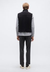 Tengerészkék ujjatlan mellény hasítottbőrből, magas gallérral, sima textúrával és minimalista dizájnnal, világosszürke ing fölött viselve.