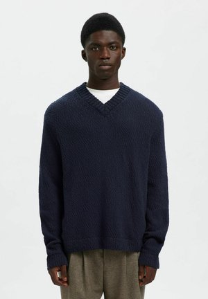 Pull à col en v bleu marine avec un motif tricoté texturé, manches longues, col côtelé et coupe décontractée. Porté par-dessus une chemise blanche.