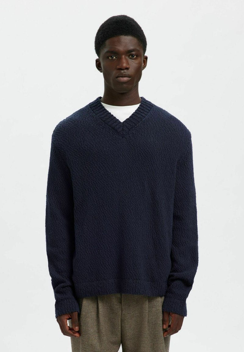 Tumši zils v-veida džemperis ar strukturētu adījumu, garām piedurknēm, ribotu apkakli un brīvu piegriezumu. Uzkopts virs baltas krekla.