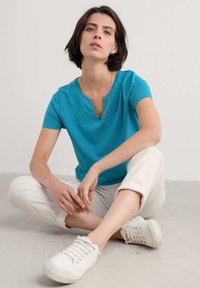 Türkisfarbenes T-Shirt mit kurzen Ärmeln und einer Kerbe am Ausschnitt, kombiniert mit beigen Caprihosen und weißen Sneakers, alles auf einem hellgrauen Boden.