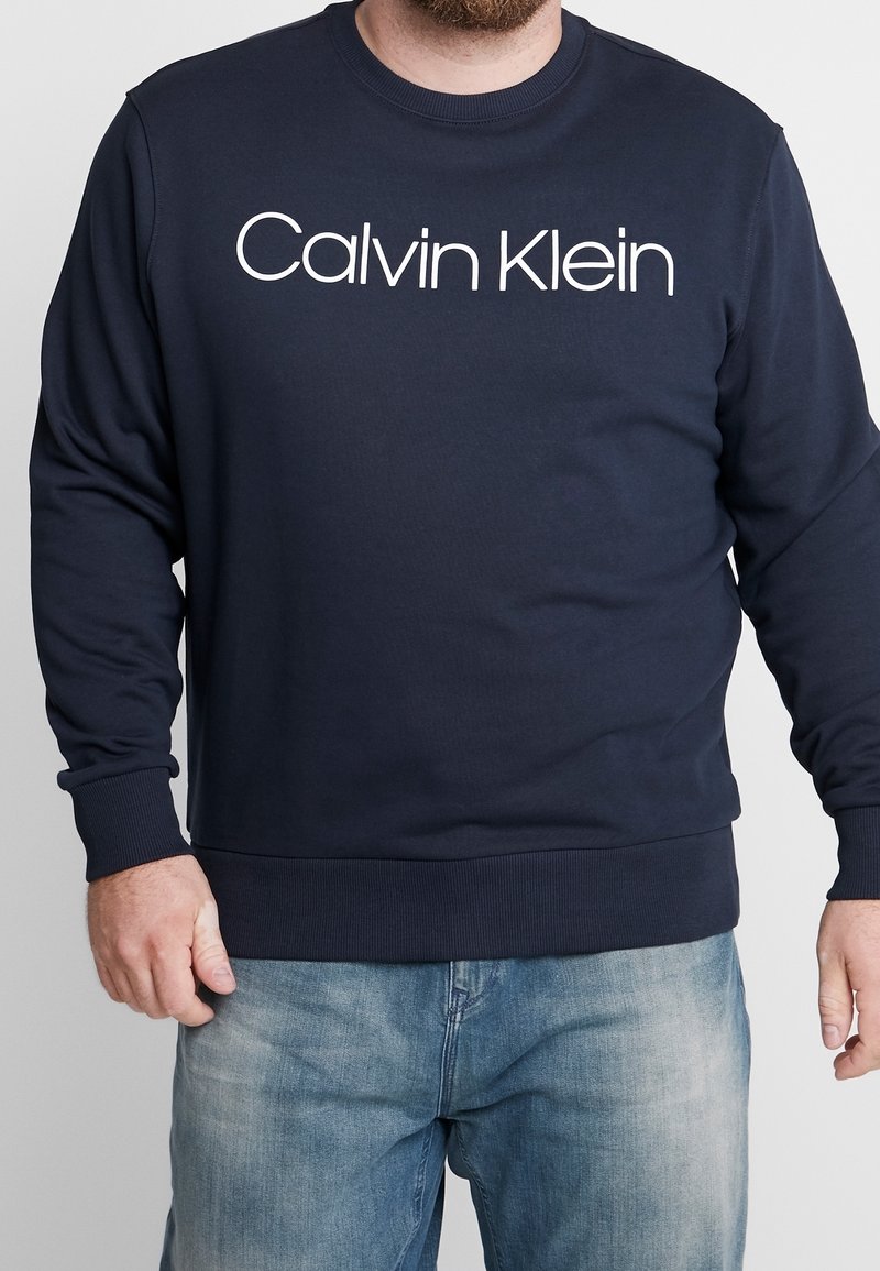 Homme portant un sweat-shirt bleu marine Calvin Klein et un jean bleu clair, debout avec les bras détendus le long du corps devant un fond uni.