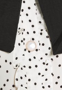 Tissu blanc à pois noirs, détails froncés, un gros bouton en perle avec un bord doré, et des bords de revers noirs d'un vêtement.