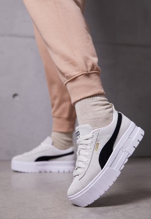 Personne portant un jogging beige et des chaussettes claires, présentant des baskets blanches Puma à plateforme avec logo noir et semelles épaisses sur un sol gris.