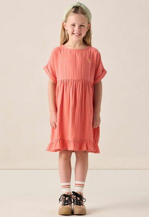Robe en coton corail avec des manches courtes, taille froncée et ourlet à volants. Associée à des baskets marron et des chaussettes blanches rayées.