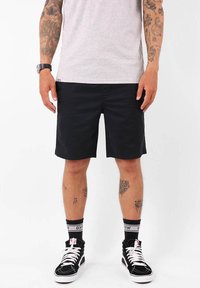 Shorts noirs en tissu lisse, à coupe droite. Portés avec des baskets montantes noires et des chaussettes à motifs.