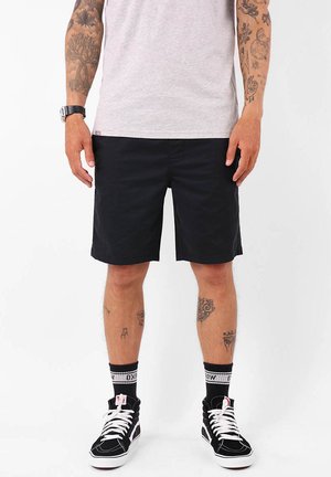 Shorts noirs en tissu lisse, à coupe droite. Portés avec des baskets montantes noires et des chaussettes à motifs.