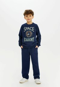 Tamsiai mėlynas džemperis su užrašu "SPACE ACADEMY", medvilnės švelnumo mišinys, laisvas fasonas; derinamas su atitinkančiomis tamsiai mėlynomis džinsinėmis kelnėmis ir baltais sportiniais batais.