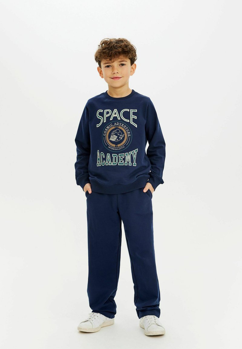 Tamsiai mėlynas džemperis su užrašu "SPACE ACADEMY", medvilnės švelnumo mišinys, laisvas fasonas; derinamas su atitinkančiomis tamsiai mėlynomis džinsinėmis kelnėmis ir baltais sportiniais batais.