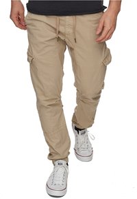Pantalons cargo beige avec une taille élastique, plusieurs poches latérales et des poignets froncés. Portés avec des baskets blanches arborant des accents noirs.