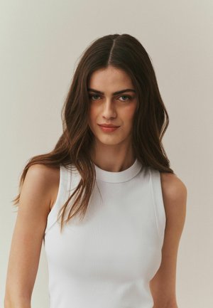 Baltas, briaunuotas, be rankovių viršus su aukštu kaklu. Dizainas prigludęs, pabrėžia švelnią tekstūrą ir subtilias kreives neutraliame fone.