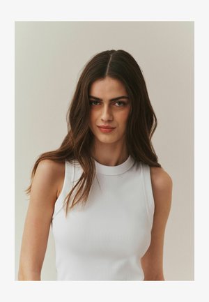 Top bianco senza maniche con collo alto e motivo a coste. Il design aderente mette in risalto una texture liscia e curve sottili su uno sfondo neutro.