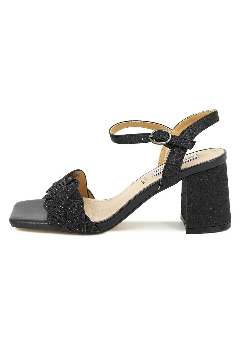 Sandali neri con tacco a spillo e punta quadrata, caratterizzati da una texture glitterata e un dettaglio a fiocco sulla parte anteriore. Una tracolla regolabile completa il design.