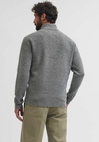 Maglione grigio lavorato a maglia con colletto alto, caratterizzato da una finitura testurizzata e polsini a costine. Indossato con pantaloni larghi di colore chiaro.