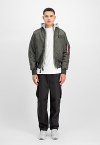 Chaqueta bomber verde oliva con capucha gris, puños elásticos y cremallera frontal. Combinada con pantalones cargo negros y zapatos gris claro.