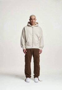 Krämfärgad zip-up hoodie med framfickor, kombinerad med bruna cargobyxor som har flera fickor, och vita sneakers. Slät textur överlag.