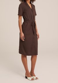 Brun klänning med kort ärm och krage, knäppning framtill och midjeband. Har två fickor och en strukturerad tyg.