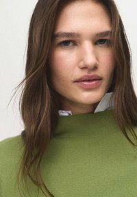 Pull en maille vert avec un col haut, présentant un tissu texturé. Une chemise blanche à col est partiellement visible en dessous. Peau lisse avec un maquillage naturel.