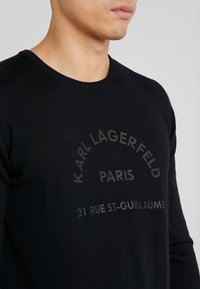 Svart stickad tröja med rundad halsringning, prydd med upphöjd text: "KARL LAGERFELD PARIS 21 RUE ST-GUILLAUME" i kontrasterande färg.