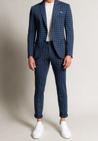 Muž v modrém slim fit obleku se vzorem psí zub, s ohrnutými kalhotami, bílým tričkem s kulatým výstřihem, kapesníčkem v kapse a bílými teniskami stojí na jednobarevném pozadí.