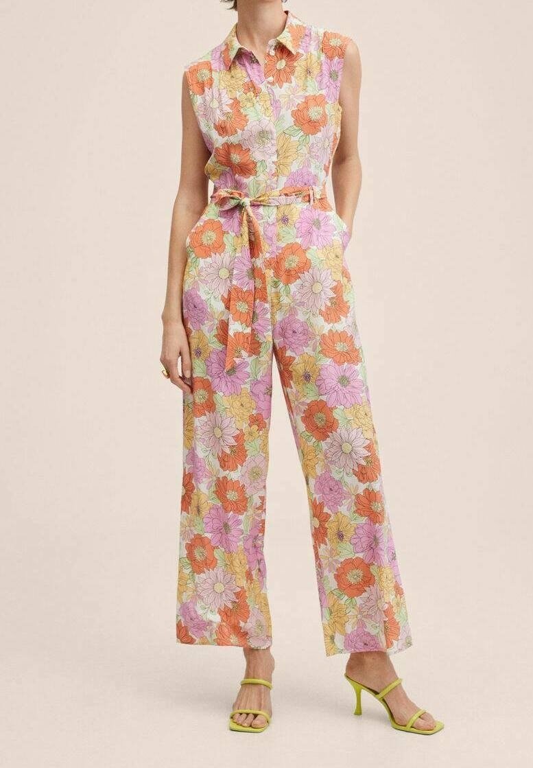 Femme portant une combinaison sans manches à fleurs orange, rose et jaune, taille ceinturée, et sandales à talons ouvertes vertes, debout.