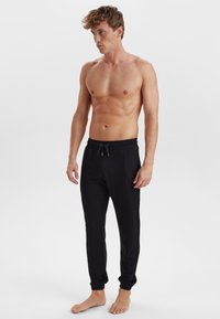 Sorte joggebukser med elastisk talje og cuffs, lavet af blødt stof, med snor. Modellen står barfodet foran en ensfarvet baggrund.