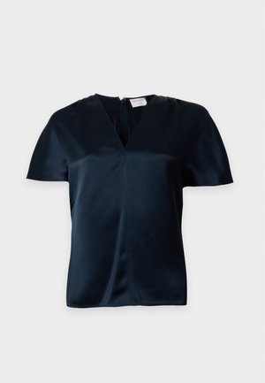 Le mannequin porte un chemisier foncé à manches courtes et à col en V, un pantalon noir à jambes larges et des bottines, sur un fond uni et clair.